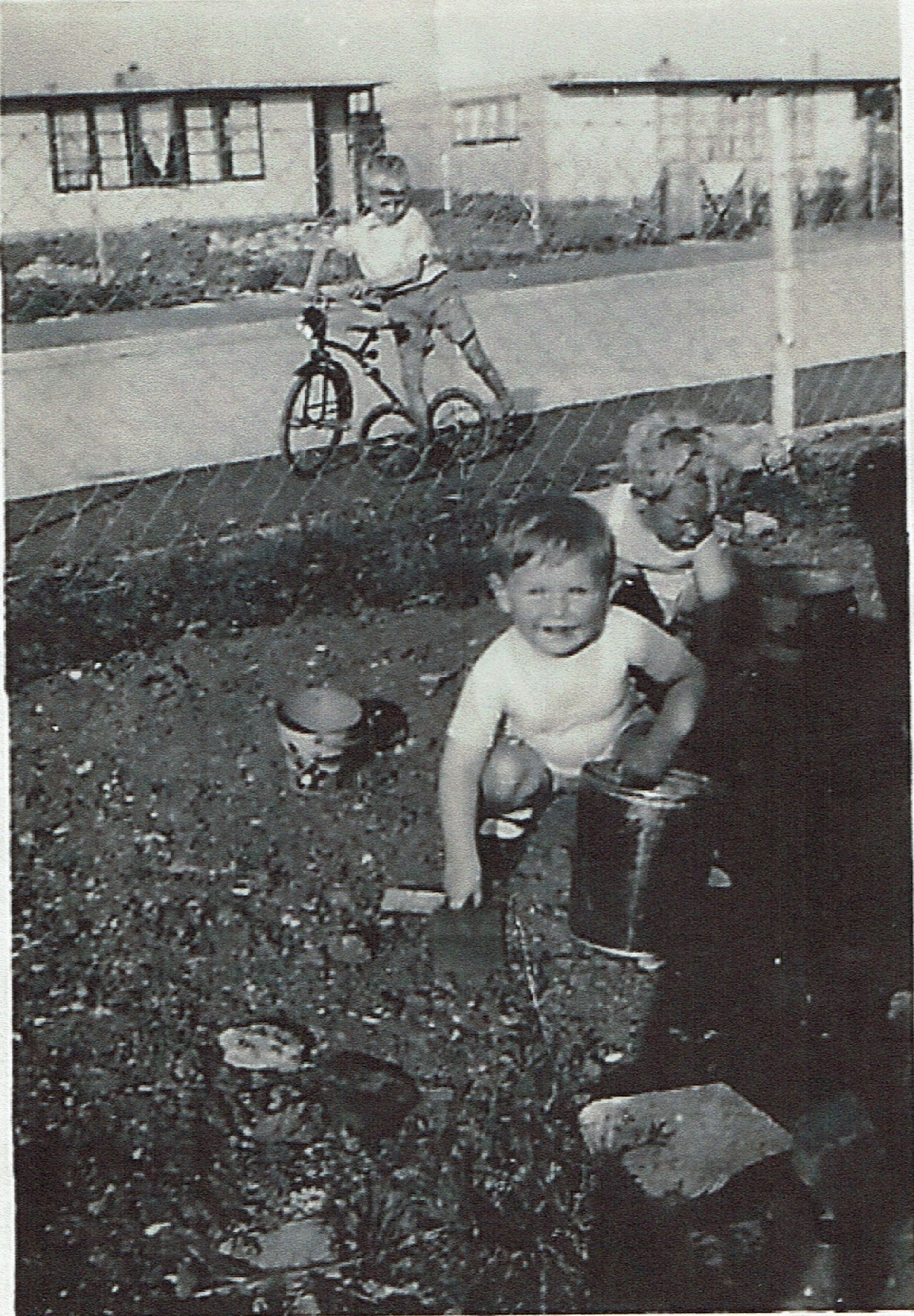 Two small children playing in the prefab garden, one in background on bike. Clement Road, Willesden