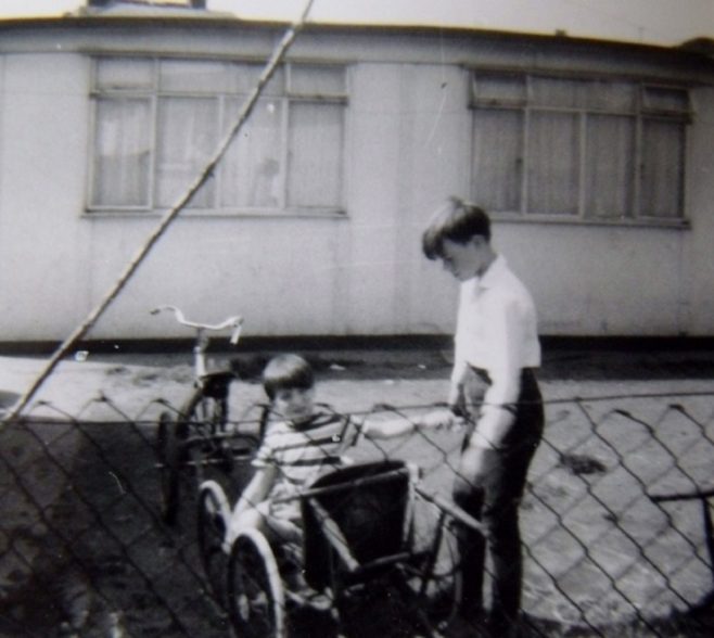 Two boys, one in a bicycle trailer, outside the prefab at Underhill Road, London SE22