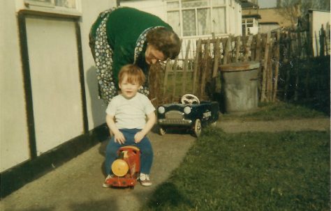 Paul sitting on a toy train, with his grandmother, in the prefab garden. Berthon Street, Deptford, London SE8