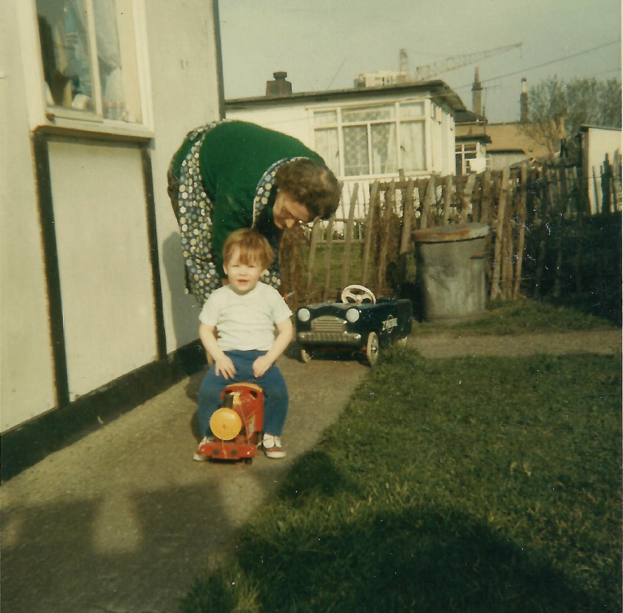 Paul sitting on a toy train, with his grandmother, in the prefab garden. Berthon Street, Deptford, London SE8